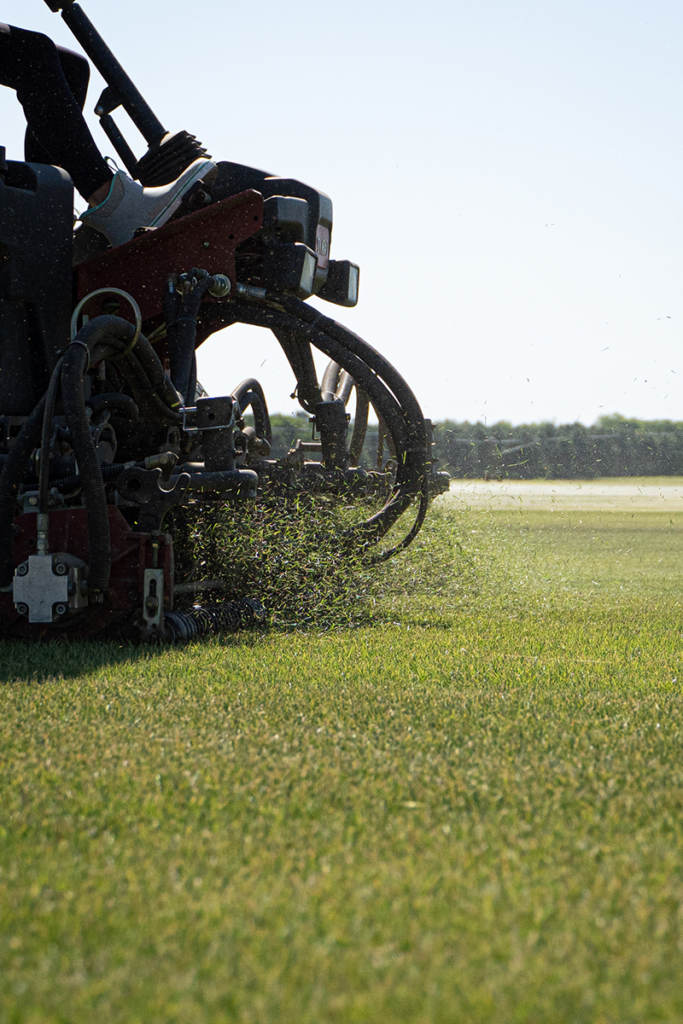 A close-up of a machine aerating a grassy field with soil spraying out in the sunlight.