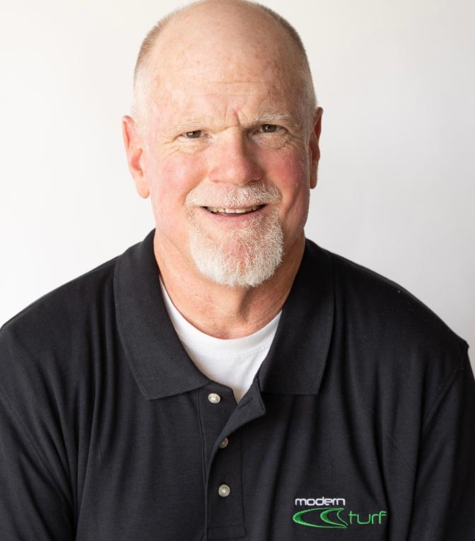 Smiling older man with a white beard wearing a black Modern Turf polo shirt in front of a plain background.