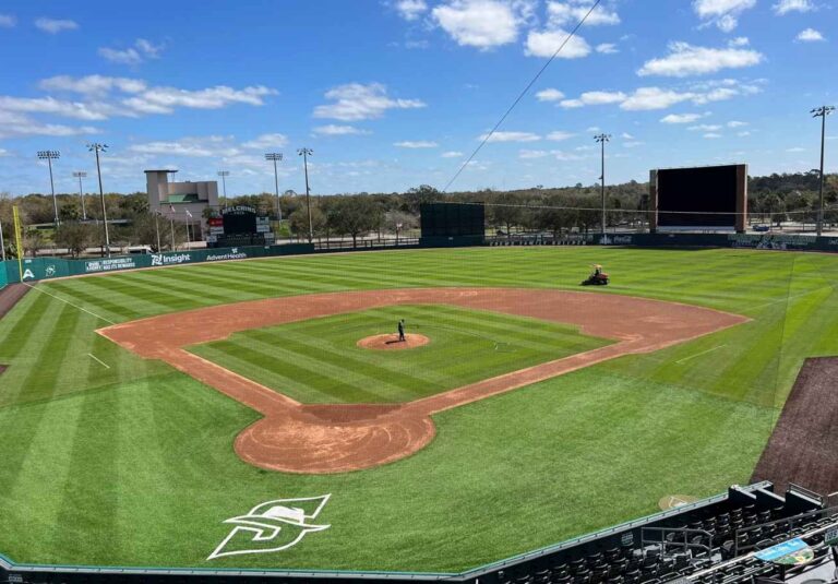 A well-maintained, empty baseball field under a blue sky with scattered clouds.