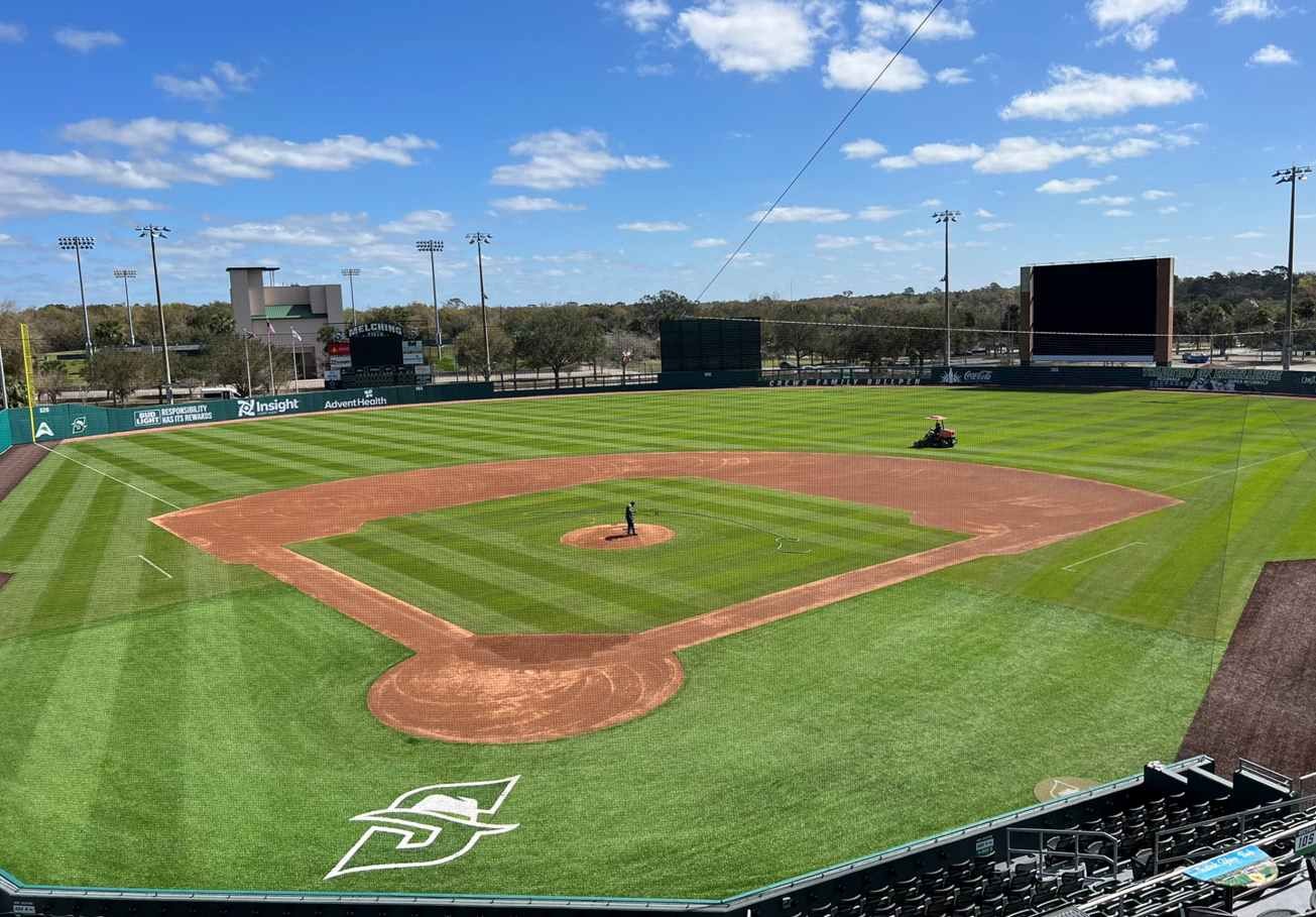 A well-maintained, empty baseball field under a blue sky with scattered clouds.